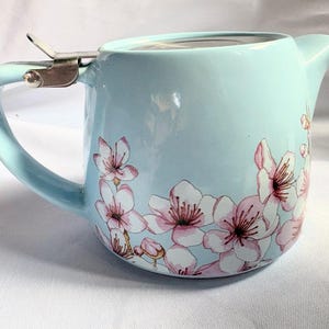 Può includere: Una teiera in ceramica azzurra con un manico curvo e un beccuccio. La teiera presenta un motivo floreale di fiori di ciliegio rosa. Un infusore in metallo è attaccato alla parte superiore.