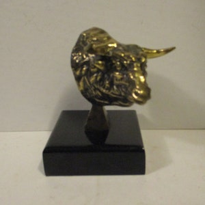 May include: A bronze-colored bull head sculpture with detailed features, mounted on a black rectangular base. The bull's horns are a lighter gold color, and the sculpture has a textured surface.