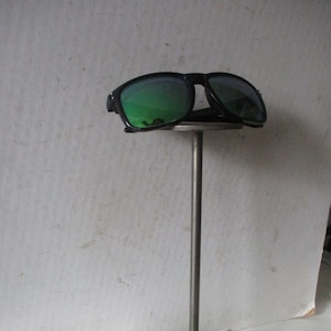 May include: A black metal sunglasses stand with a round base and a tall, slender pole. A pair of black sunglasses with green lenses are displayed on the stand.