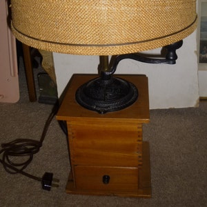 May include: A vintage coffee grinder lamp with a wooden base and a burlap lampshade. The lamp has a black metal arm and a black metal grinder.