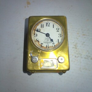 May include: A gold-colored clock with a white face and black hands. The clock has a round face with Roman numerals and a small, round window at the bottom. The clock is mounted on a rectangular base with two knobs on the side.