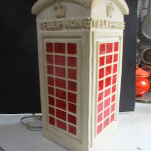 May include: A cream-colored telephone booth lamp with red window panes. The lamp has the word "TELEPHONE" on the top and a crown emblem. The lamp is a decorative item.