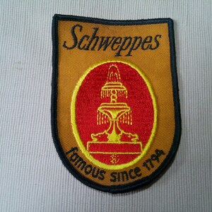 May include: Embroidered patch featuring the Schweppes logo. The patch has a shield shape with a gold background and a black border. The logo includes the word "Schweppes" in black script and a red oval with a fountain design.