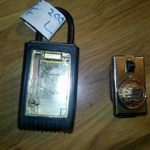 May include: A black key safe with a silver interior and a black handle is next to a small, gold-colored combination lock. A piece of paper with blue writing is attached to the key safe.