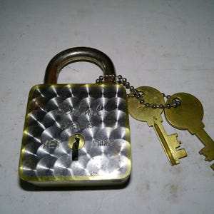 May include: A brass padlock with a silver-toned face featuring a circular engine-turned pattern. The lock is engraved with the text Sentinel, 5 Levers, and Key Proof. Two brass keys are attached to the shackle by a small metal ball chain.