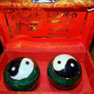 May include: Two spherical exercise balls with green, black, and white yin-yang symbols, housed in a red fabric-lined box. The box lid features gold text reading Directions Healthy Ball and Made in China with a decorative gold graphic.