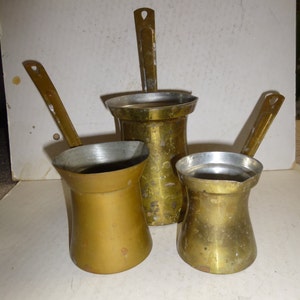 May include: Three brass coffee pots of different sizes with handles. The pots are all tarnished and have a worn look.