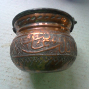 May include: A small, antique copper pot with a rounded body and a dark rim. The pot features embossed Arabic script and decorative patterns around the base. The copper has a weathered, aged appearance.