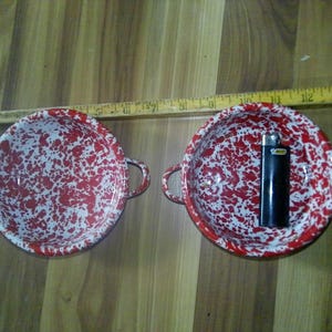May include: Two round, white enamel bowls with red speckled patterns and two side handles. A yellow wooden ruler is positioned above the bowls, showing a length of approximately 14 inches. A black Bic lighter is placed inside the right bowl.