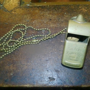May include: A vintage brass whistle with a ball chain, labeled "The Acme Thunderer, Made in England." The whistle is rectangular with a rounded top and a small opening. The chain is made of small, round beads.