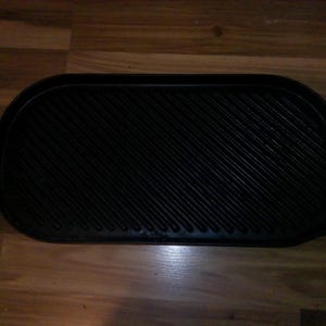May include: Black cast iron griddle with a ribbed cooking surface and two handles. The griddle is oval-shaped and sits on a wooden surface. Ideal for grilling meats and vegetables.