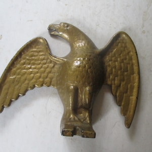 May include: A cast metal eagle sculpture with outstretched wings, finished in a gold color. The eagle is depicted in a standing pose with detailed feather textures on its wings. The sculpture is likely a decorative item.