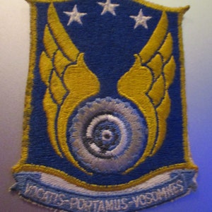 May include: A blue and yellow embroidered patch with a white spiral design and three white stars. The text "VINCIT QUI PATITUR - VOS SUMUS" is on the bottom of the patch.