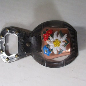 May include: A metal bottle opener with a copper cowbell attached. The cowbell is decorated with painted flowers in red, white, blue, and yellow.