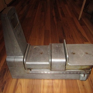 May include: A vintage silver-colored postal scale with three weighing platforms and a graduated scale. The scale has a black label with the word "POSTAGE" printed on it.