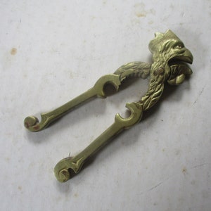 May include: A gold-colored metal nutcracker shaped like a bird with a detailed design. The nutcracker has a hinged jaw and a lever to crack nuts.