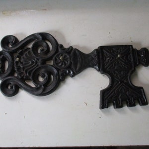 May include: Ornate black cast iron key-shaped wall decor. The key features intricate scrollwork and a detailed handle. This decorative piece is likely used for interior design or as a unique wall hanging.