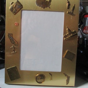 May include: A gold-colored metal picture frame with travel-themed embellishments. The frame features miniature depictions of landmarks, a map of the United States, and other travel-related icons. The frame is designed to hold a photograph.