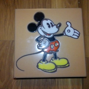 May include: Square ceramic tile featuring a cartoon image of Mickey Mouse. The character is depicted in his classic outfit with black ears, a white face, red shorts, yellow shoes, and white gloves. The tile has a light brown background.