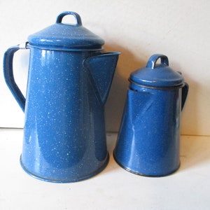 May include: Two blue enamelware coffee pots with white speckles. The larger pot has a spout and handle, while the smaller one is a sugar bowl. Both have lids with loop handles. A classic camping or kitchen set.