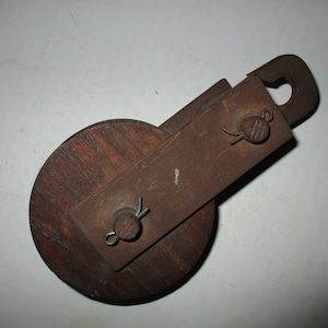May include: A rusty metal and wooden pulley with two metal hooks and a wooden wheel. The pulley is designed for lifting heavy objects.
