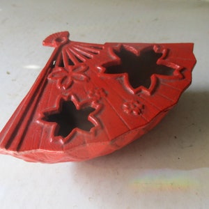 Puede incluir: Un plato de cerámica rojo en forma de abanico con tres diseños de hojas de arce recortados. El plato tiene un borde elevado y una superficie texturizada.