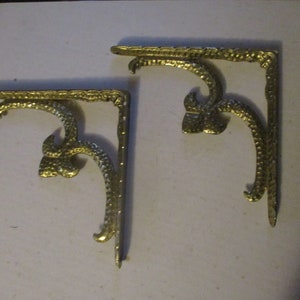 May include: Two gold metal corner brackets with a decorative scroll design. The brackets are about 4 inches long and 3 inches wide.