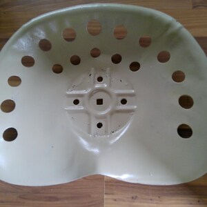 May include: An off-white tractor seat with multiple circular holes for ventilation. The seat has a curved shape and a central mounting area with a square indentation. The seat is made of metal and has a slightly worn appearance.
