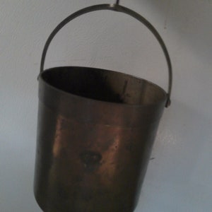 May include: A copper bucket with a metal handle. The bucket is empty and has a shiny, slightly tarnished surface.