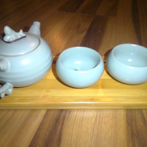 May include: A light blue ceramic tea set with a teapot and two matching cups, presented on a rectangular bamboo tray. The teapot has a curved handle and a decorative lid. The cups are round.