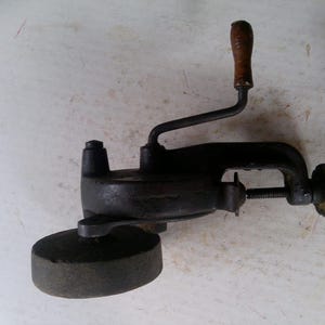 May include: A vintage, cast iron hand-cranked grinder with a wooden handle. The grinder is black with a circular grinding stone and a clamp for securing it to a surface. The crank is attached to the top of the grinder.