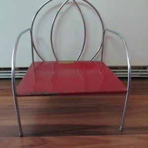 May include: A small, vintage red chair with a metal frame. The chair has a rectangular seat and a curved backrest made of silver-colored metal. The chair is on a wooden floor.