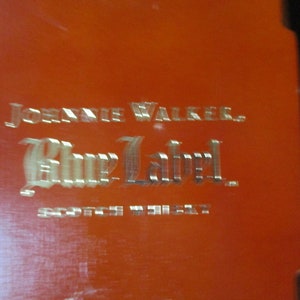 Pu&ograve; includere: Primo piano di una superficie arancione con le parole "JOHNNIE WALKER. Blue Label. SCOTCH WHISKY" in un carattere metallico in rilievo. Il testo &egrave; centrato e occupa la maggior parte dell'inquadratura.
