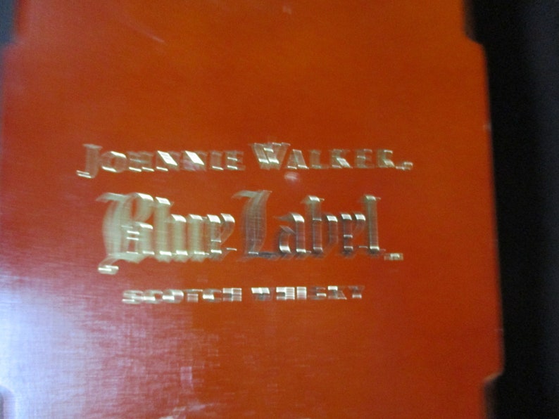 Pu&ograve; includere: Primo piano di una superficie arancione con le parole "JOHNNIE WALKER. Blue Label. SCOTCH WHISKY" in un carattere metallico in rilievo. Il testo &egrave; centrato e occupa la maggior parte dell'inquadratura.