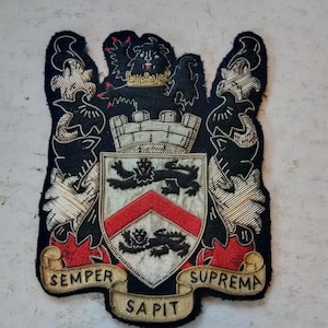 May include: A black and gold embroidered crest with a lion, a castle, and a red and white shield. The crest is surrounded by a gold scroll with the words "SEMPER SAPIT SUPREMA".