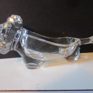 May include: A clear glass ashtray in the shape of a dog. The ashtray has a rectangular bowl and a tail. The dog's head is detailed with ears and a snout. The ashtray is sitting on a white surface.