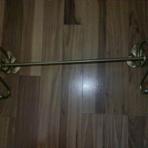 May include: A brass-toned metal towel bar featuring a horizontal rod supported by two curved brackets. Each bracket is accented with a decorative leaf-shaped mounting plate and terminates in a small hook at the bottom for additional storage.