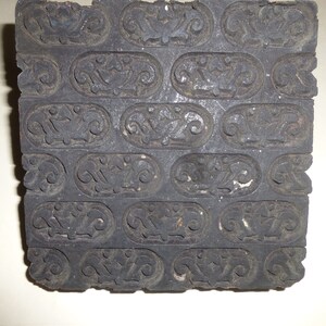 May include: A dark gray, rectangular wooden printing block with a repeating pattern of oval shapes containing floral and bird-like designs. The block has a worn, aged appearance, suggesting it is vintage or antique.