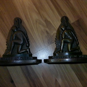 May include: A pair of bronze-toned metal bookends featuring a kneeling figure in a recessed archway. Each base is inscribed with the text University of Toledo. The design includes a torch emblem at the top of the arch and a gear-like border.