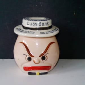 May include: A vintage ceramic money bank shaped like a grumpy face with a hat that reads "Cuss Bank". The bank has a red mouth, brown eyebrows, and a black and yellow detail at the bottom. The bank is a novelty item.