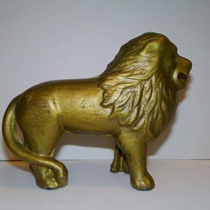 May include: A gold-colored cast iron lion figurine. The lion is standing on all four legs with its head held high. The lion has a long mane and a detailed face.