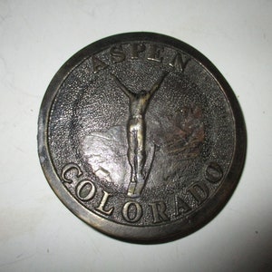 May include: A round, bronze-colored belt buckle with the words "Aspen Colorado" and a skier jumping in the air.