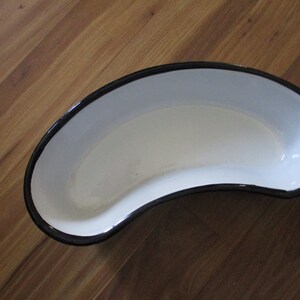 May include: White kidney-shaped enamelware basin with a black rim. The basin is empty and sits on a wooden surface. This medical or laboratory supply is a classic design.