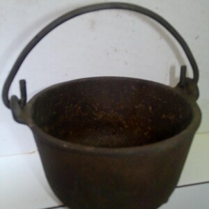 May include: A dark brown cast iron cauldron with a curved metal handle. The cauldron has a rounded bowl shape and small feet. The handle is attached to the sides of the cauldron with small metal loops. The interior of the cauldron appears to be textured.