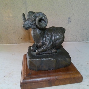 May include: A bronze sculpture of a ram resting on a stone base, mounted on a wooden plinth. The ram has large, curved horns and detailed fur texture. The sculpture is a dark bronze color.