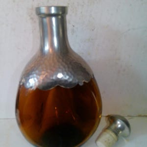 May include: An amber-colored glass decanter with a silver-colored metal top and a matching stopper. The decanter has a rounded body and a narrow neck. The stopper is lying next to the decanter.