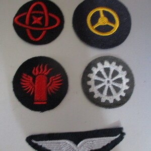 May include: Five embroidered patches on a white background. The patches are circular with black fabric and various colored embroidery. The patches feature a red circle with a red star inside a red ring, a yellow steering wheel, a red flame with wings, a white gear, and a white wing.