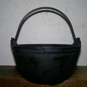May include: A black cast iron pot with a rounded base and a thick, curved metal handle attached to two side lugs. The exterior surface of the pot has a textured, matte finish, and the rim is smooth and circular, showing a sturdy construction.