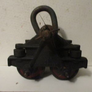 May include: A vintage black metal pulley system with a curved handle and two red-rimmed wheels. The pulley is designed for heavy-duty use, with a sturdy construction and a weathered appearance. The design suggests it is an industrial or antique item.