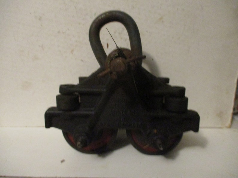 May include: A vintage black metal pulley system with a curved handle and two red-rimmed wheels. The pulley is designed for heavy-duty use, with a sturdy construction and a weathered appearance. The design suggests it is an industrial or antique item.